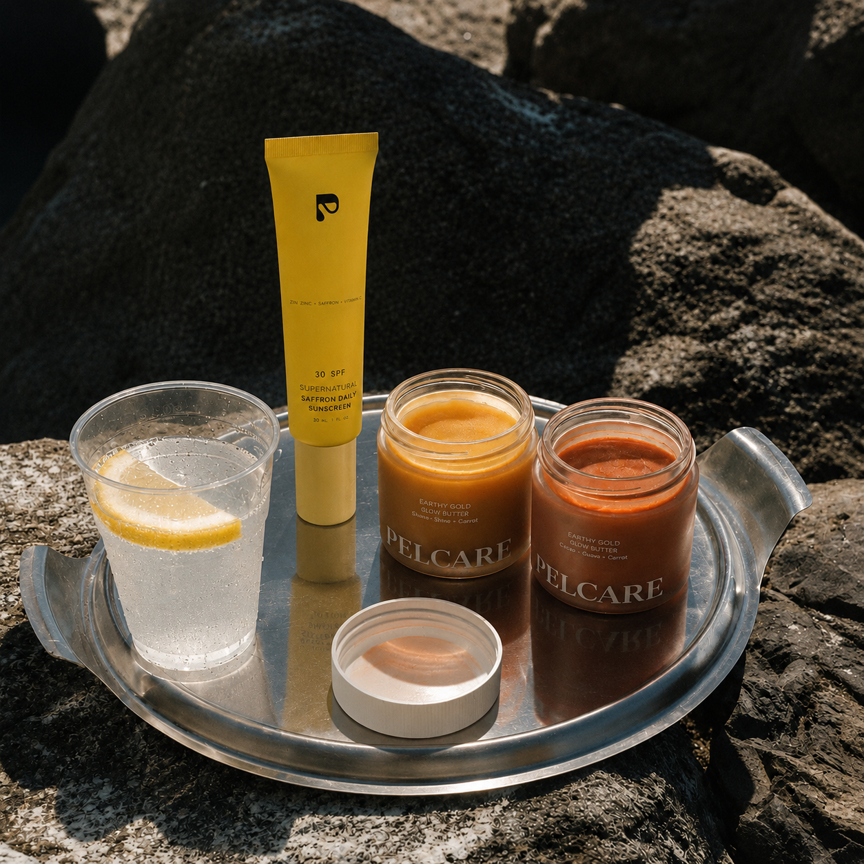 Pelcare summer skincare products with SPF saffron daily sunscreen and glow butter jars on a silver tray, styled on sunlit black rocks with a refreshing lemon drink.
